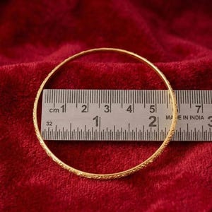 May include: A gold bangle with a detailed pattern, set against a deep red background. The bangle measures approximately 6 cm in diameter, as indicated by the ruler. The circular piece of jewellery is likely made of metal.