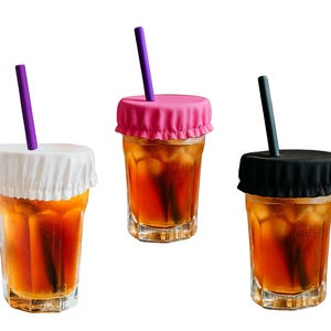 May include: Three iced tea glasses with colorful lids and straws. One glass has a white lid and purple straw, another has a pink lid and purple straw, and the third has a black lid and black straw. The glasses are filled with iced tea and ice cubes.