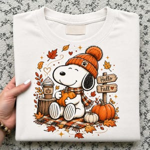 Puede incluir: Una sudadera blanca de cuello redondo con un gráfico de un perro de dibujos animados con gorro y bufanda de punto naranja, sosteniendo una calabaza. El diseño incluye una taza de café, hojas de otoño y un letrero con el texto Hello Fall.
