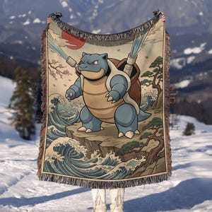 May include: A woven throw blanket featuring a blue turtle-like creature with two cannons on its back, spraying water. The design includes stylized ocean waves, a red sun, cherry blossoms, and a pine tree on a cliffside, finished with fringed edges.