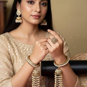 May include: A set of gold-toned bangles featuring white beaded accents and dangling bell-shaped charms with pearl-like fringe, paired with matching gold-toned drop earrings and a circular ring with a central stone and surrounding bead detail.