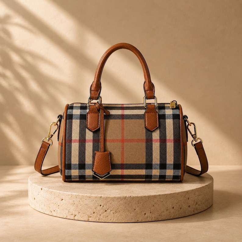 May include: A tan and plaid handbag with brown leather handles and trim. The bag features a classic plaid pattern in tan, black, red, and white. It has a top handle and a detachable shoulder strap. The bag is displayed on a round, stone-colored pedestal.