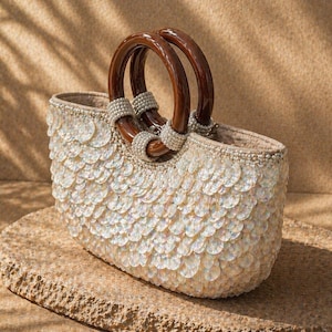 May include: An oval-shaped handbag with a white sequined exterior and brown wooden handles. The handles are connected to the bag with pearl-like beads. The bag is sitting on a stone surface.