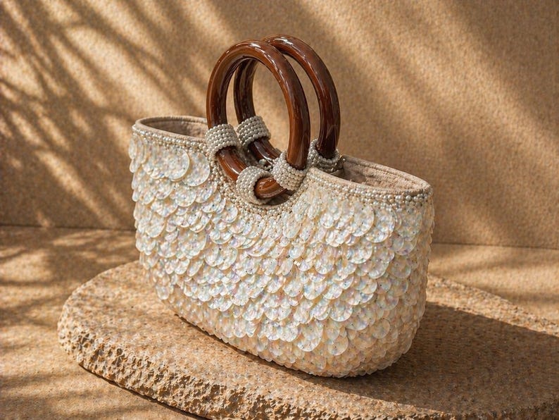 May include: An oval-shaped handbag with a white sequined exterior and brown wooden handles. The handles are connected to the bag with pearl-like beads. The bag is sitting on a stone surface.