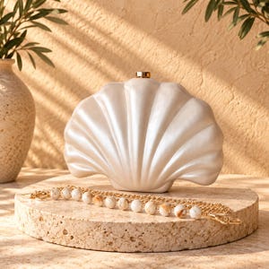 Peut inclure: Une pochette en forme de coquillage blanc nacré avec un fermoir en métal doré sur le dessus. Le sac comprend une bandoulière amovible composée d'une chaîne en métal doré alternée avec des perles rondes marbrées blanches et fauves.