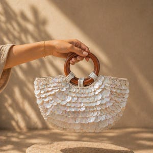 May include: A white, shell-covered handbag with a wooden handle. The bag is held by a person. The handle is a dark brown color and is attached to the bag with white beads. The bag is a half-circle shape.