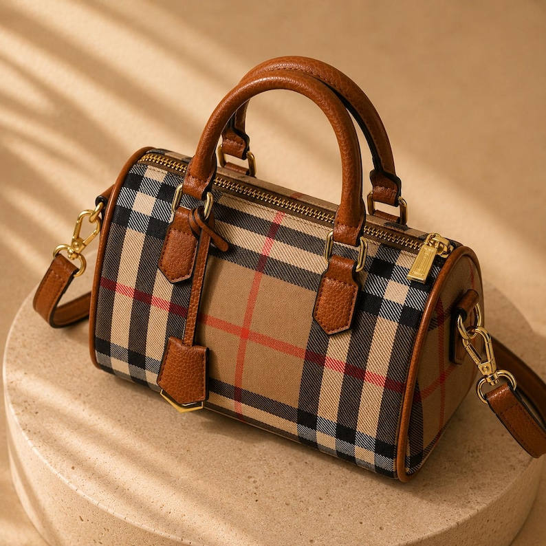 Puede incluir: Bolso con forma de barril a cuadros beige, negro y rojo, con asas y ribetes de cuero marr&oacute;n. El bolso tiene una cremallera dorada y una correa para el hombro desmontable. El bolso est&aacute; sobre una superficie clara.