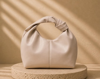 May include: A beige, small handbag with a knotted handle. The bag is made of a soft material and has a curved shape. It sits on a round, textured, beige pedestal. The background is a solid beige color with shadows.