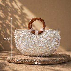 May include: A white, shell-covered handbag with a wooden handle. The bag is approximately 10 inches tall and 12 inches wide. The handle is a dark brown, circular shape. The bag is sitting on a stone surface.