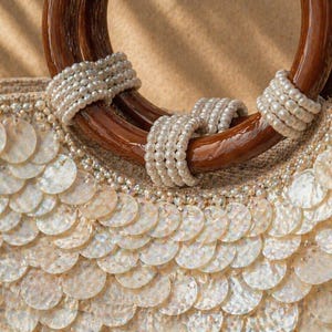 May include: Close-up of a beige handbag with a wooden handle. The handle is a dark brown color and is wrapped with white pearls. The body of the bag is covered in white, round, shell-like embellishments.