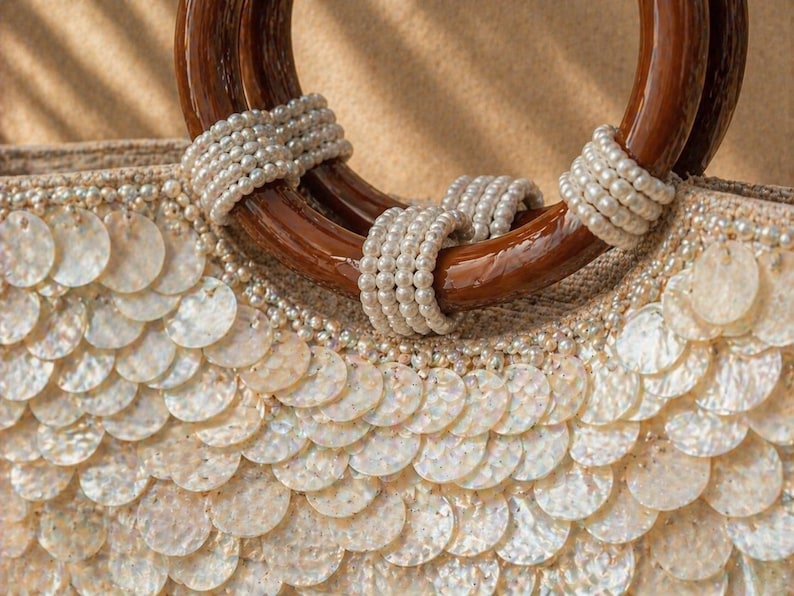 May include: Close-up of a beige handbag with a wooden handle. The handle is a dark brown color and is wrapped with white pearls. The body of the bag is covered in white, round, shell-like embellishments.