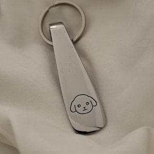 May include: A silver-toned metal keychain featuring a flat, elongated, tapered pendant with a rounded bottom edge. The surface of the pendant is engraved with a simple, black-lined graphic of a dog's face, including ears, eyes, and a nose.