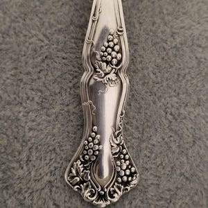 May include: A silver-toned metal pendant crafted from the handle of a spoon, featuring an embossed design of grape clusters and vine leaves. The pendant is suspended from a silver-colored ball chain necklace, showcasing a polished finish.