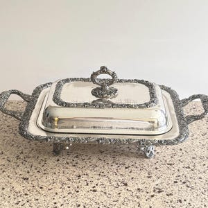 May include: Silver-plated serving dish with a rectangular shape and a lid. The dish features ornate detailing around the edges and handles, and a decorative handle on the lid. The dish is elevated on small feet.