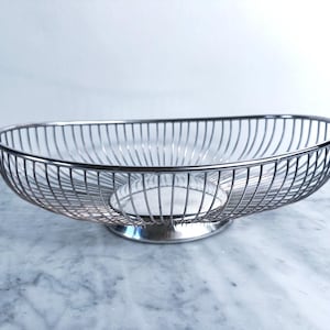 May include: A silver wire fruit bowl, oval in shape. The bowl has a rounded base and vertical wires forming the sides. The bowl is empty and rests on a marble surface. The dimensions are approximately 30 cm in length.