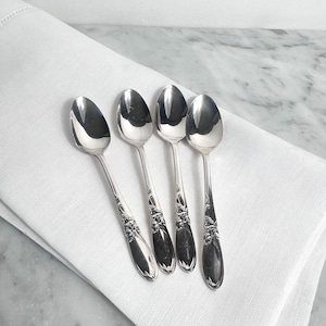 May include: Four silver spoons with ornate floral detailing on the handles are arranged on a white linen napkin. The spoons have a reflective surface, and the napkin is neatly folded. The background is a marble surface.