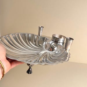 May include: A silver-toned, shell-shaped serving dish with three small cup holders and a decorative foot. The dish has a fluted design and is held by a hand with a gold bracelet.
