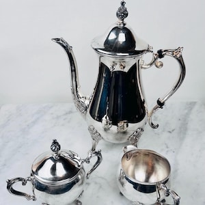 May include: A silver tea set with a coffee pot, sugar bowl, and creamer. The coffee pot has a tall, ornate design with a curved spout and a decorative handle. The sugar bowl and creamer have matching designs. The set is displayed on a marble surface.