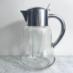 May include: A clear glass pitcher with a stainless steel lid, handle, and spout. The pitcher has a cylindrical ice tube in the centre. The handle is curved. The pitcher sits on a marble surface.