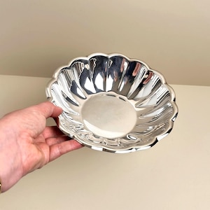 May include: A shiny silver bowl with a scalloped edge. The bowl has a smooth, reflective surface and a shallow, rounded shape. The bowl is held by a hand.