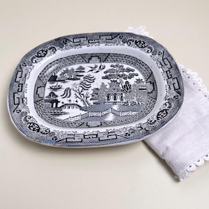 May include: Oval blue and white ceramic serving platter with a Willow pattern design. The platter is approximately 14 inches long and rests on a white, scalloped-edge linen napkin.