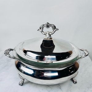 May include: A silver-plated serving dish with a lid and ornate handles. The dish has a clear glass insert and sits on three decorative feet. The lid features a detailed handle, and the piece reflects light, suggesting a polished finish.