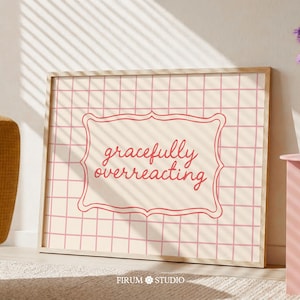 Puede incluir: Un cuadro enmarcado con las palabras "gracefully overreacting" en un marco decorativo. El fondo es un patrón de cuadrícula en color rosa claro. El marco es de color madera clara. El cuadro está apoyado contra una pared.