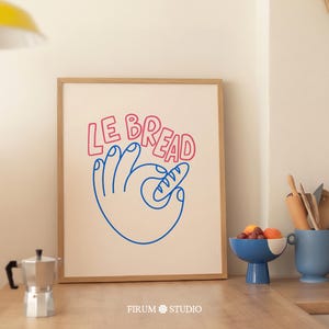 Puede incluir: Una impresión artística enmarcada con las palabras "LE BREAD" en rojo y azul. La obra de arte representa una mano sosteniendo una barra de pan. Un cuenco azul con fruta y un soporte de cerámica azul para utensilios están sobre una superficie de madera.