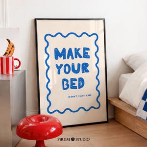 Könnte beinhalten: Gerahmtes Poster mit dem Text "MAKE YOUR BED" in Blau, umgeben von einem wellenförmigen blauen Rand. Ein roter Hocker und eine gestreifte Tasse sind ebenfalls zu sehen. Das Poster befindet sich in einem Raum mit einem Bett und einem Bananenbild.