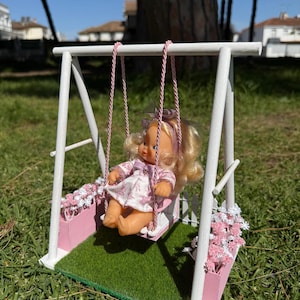Puede incluir: Una muñeca en miniatura en un columpio de madera blanco con detalles rosas y una base verde. La muñeca tiene cabello rubio y lleva un vestido rosa. El columpio tiene cuerdas rosas y está decorado con flores rosas y blancas.