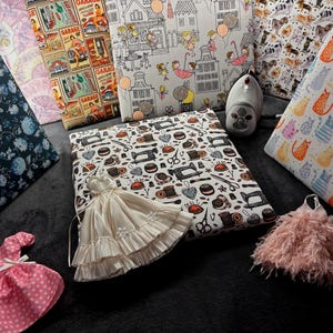 Puede incluir: Una colección de telas estampadas, que incluyen estampados florales, letreros de garaje vintage y gatos. Se exhiben vestidos en miniatura en rosa y crema con una plancha. Las telas son adecuadas para proyectos de costura y manualidades.