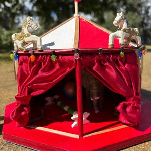 May include: A miniature circus tent with a red and white striped roof, red curtains, and a red base. Two white horses with gold accents sit atop the tent. The tent has a small red flag and decorative lights.