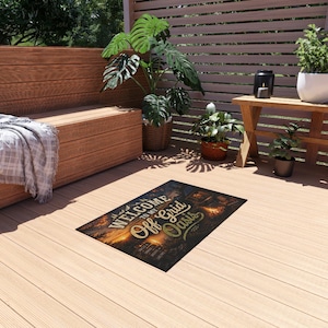 Può includere: Uno zerbino rettangolare raffigurante una scena boschiva al tramonto con un falò e il testo "WELCOME TO MY Off Grid Oasis" in un carattere stilizzato. Il tappetino presenta uno sfondo scuro con calde tonalità arancioni e gialle.