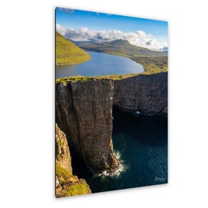May include: A scenic landscape photograph featuring a lake and a cliff. The lake is surrounded by green hills and the cliff is a dark brown color. The sky is blue with some clouds. The water is a deep blue color.