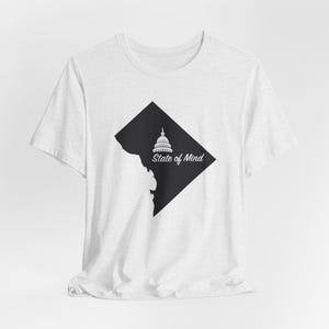 May include: A white t-shirt featuring a black graphic of the Washington D.C. outline and the Capitol building. The words "State of Mind" are printed below the Capitol building in a white, stylized font. The shirt is made of a soft, light-colored fabric.
