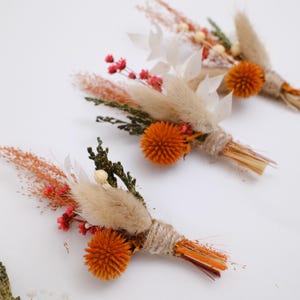 May include: Dried flower boutonnieres with orange, beige, and red hues. Each boutonniere features an orange globe flower, surrounded by dried grasses and flowers, bound with twine. The background is plain white.