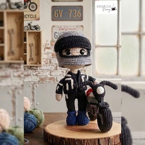 Może przedstawiać: Szydełkowa lalka amigurumi w szaro-czarnym kasku motocyklowym, czarnej kurtce z białymi akcentami i niebieskich butach, stojąca obok małego czarnego szydełkowego motocykla na okrągłej drewnianej podstawie.