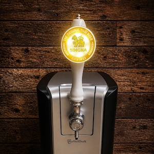 May include: A silver and black PerfectDraft beer dispenser with a tap and a glowing Singha beer logo. The dispenser has a digital temperature display showing 3 degrees. The background is a wooden surface.