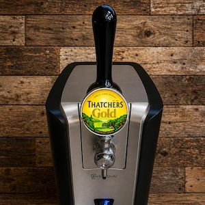 May include: A black and silver Perfect Draft beer dispenser with a tap handle. The dispenser features a Thatchers Gold cider label and a digital temperature display. The unit is set against a wooden background.
