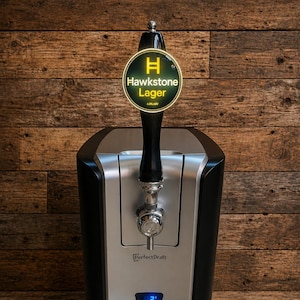 May include: A black and silver PerfectDraft beer dispenser with a Hawkstone Lager tap. The tap handle has a circular sign with the Hawkstone Lager logo. The dispenser has a digital display and a drip tray.