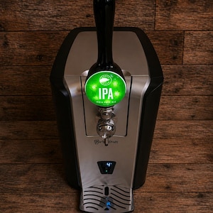 May include: A black and silver Perfect Draft beer dispenser with a tap handle and a green IPA label. The dispenser has a digital display showing the temperature and is set against a wooden background.