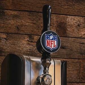 May include: A black and silver PerfectDraft beer dispenser with an NFL logo on the tap handle. The dispenser has a digital temperature display showing 8.4 degrees. The unit sits on a wooden surface.