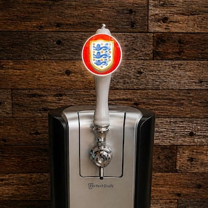 May include: A black and silver PerfectDraft beer dispenser with a tap featuring the England national team emblem. The dispenser has a digital temperature display and a drip tray. The background is a wooden wall.