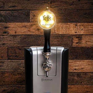 May include: A black and silver PerfectDraft beer dispenser with a tap featuring a Leffe Blonde logo. The dispenser has a digital display showing a temperature of 83 degrees. The background is a wooden wall.