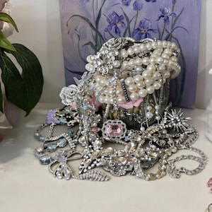 Może przedstawiać: Kolekcja biżuterii w stylu vintage wylewa się z ozdobnego srebrnego pojemnika. Asortyment obejmuje perłowe naszyjniki, bransoletki z cyrkoniami i broszki z różowymi i przezroczystymi kamieniami szlachetnymi. W tle obraz fioletowych irysów.