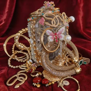 May include: A display of gold jewellery, including bracelets, rings, and necklaces, overflowing from a glass jar. The jewellery features pearls, gemstones, and decorative elements like a butterfly and a snake. The background is red velvet.