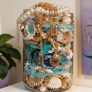May include: A clear glass jar overflowing with jewelry and seashells. Gold chains, pearl necklaces, and turquoise beads mix with various shell shapes. The jar is filled with a variety of textures and colors, creating a coastal theme.
