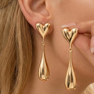 Könnte beinhalten: Goldfarbene herzförmige Ohrhänger. Die Ohrringe haben einen herzförmigen Stecker und einen tropfenförmigen Anhänger. Die Ohrringe sind aus glänzendem, reflektierendem Metall gefertigt. Die Ohrringe werden von einer Person getragen.