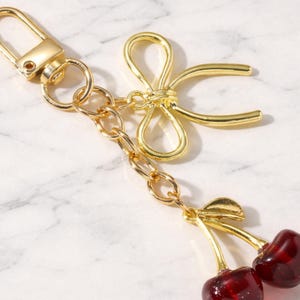 May include: Gold-toned keychain featuring a bow and a pair of cherries. The cherries are a deep red color with gold stems. The keychain includes a clasp and a chain, all in a shiny gold finish.