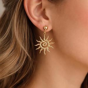 Op de afbeelding: Goudkleurige oorbellen met een zonnemotief. Elke oorbel heeft een kleine, ronde knop aan de bovenkant, verbonden met een gestileerd zonontwerp met stralende stralen en een centrale spiraal. De oorbellen worden op het oor van een persoon getoond.